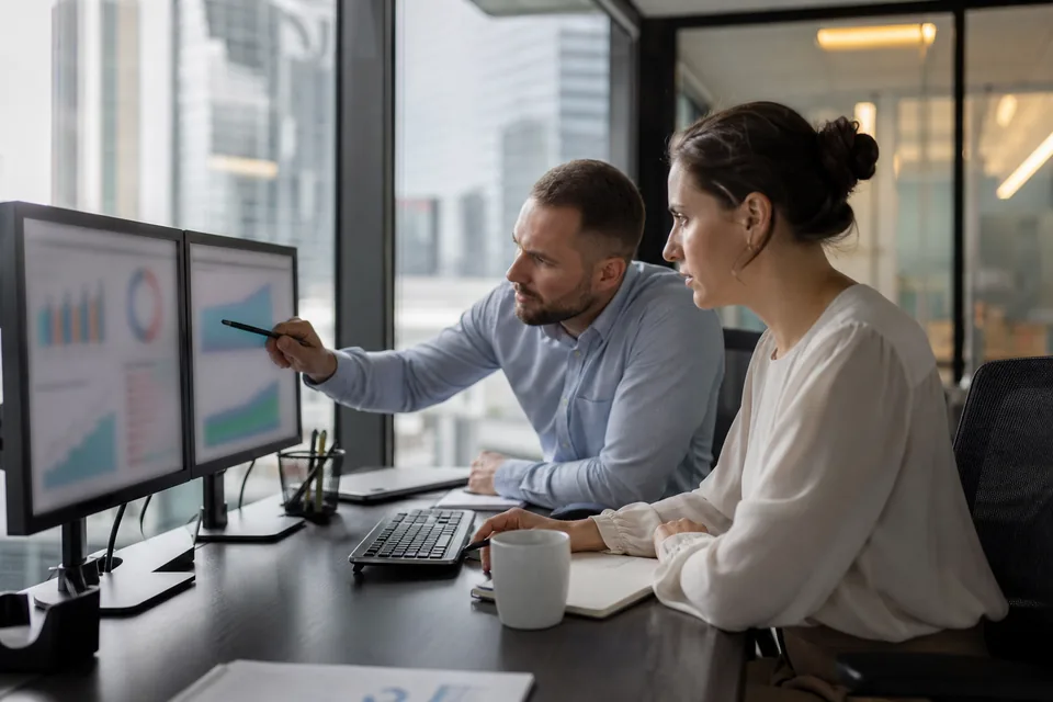 Portfolio finance lead and PMO manager at a bank reviewing initiative budgets against actuals on two monitors