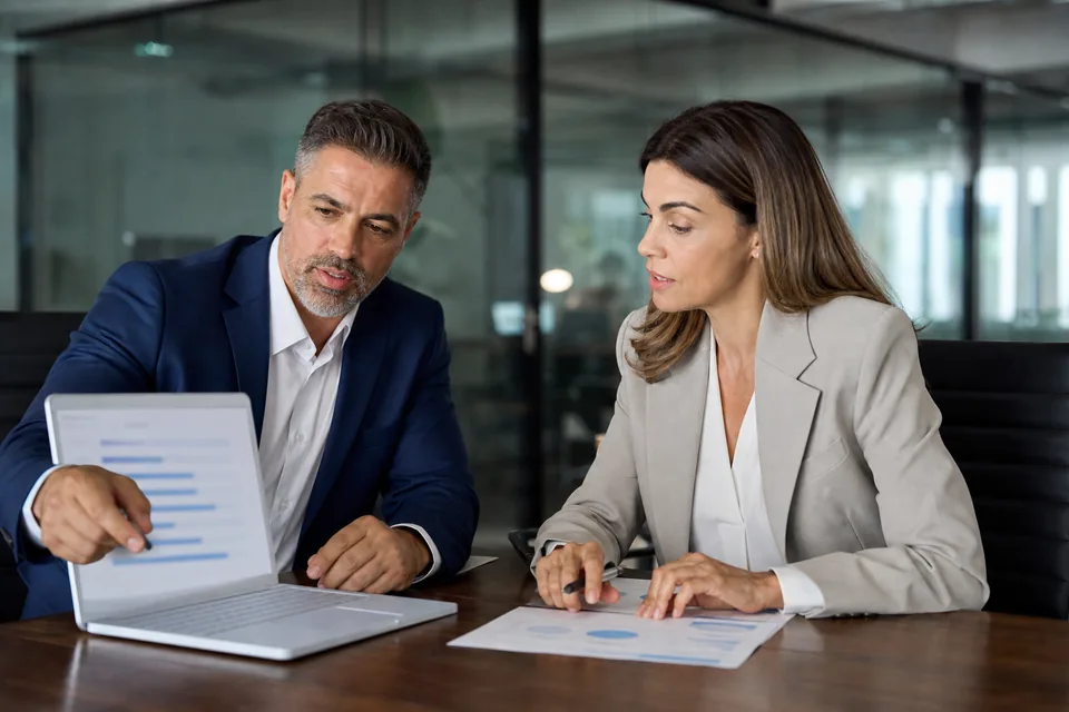 PMO director and IT finance manager comparing yearly software cost bars on a laptop in a glass-walled office